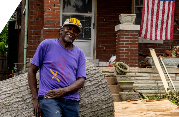 A smiling man in a purple shirt leaning against a large fallen tree trunk in front of a brick house with an American flag.
