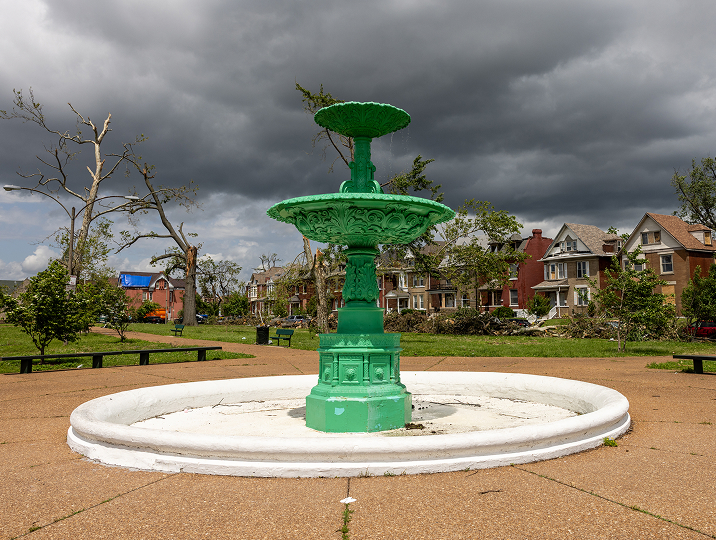 A bright green tiered fountain in a St. Louis park with storm-damaged trees and houses under a dark, cloudy sky.