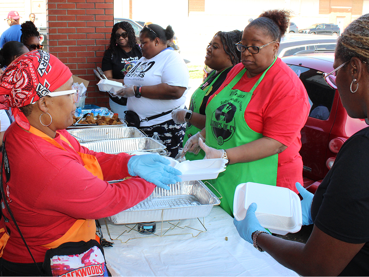 Volunteers in red shirts and green aprons serve meals into takeout containers at an outdoor community event.