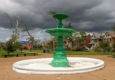 A bright green tiered fountain in a St. Louis park with storm-damaged trees and houses under a dark, cloudy sky.